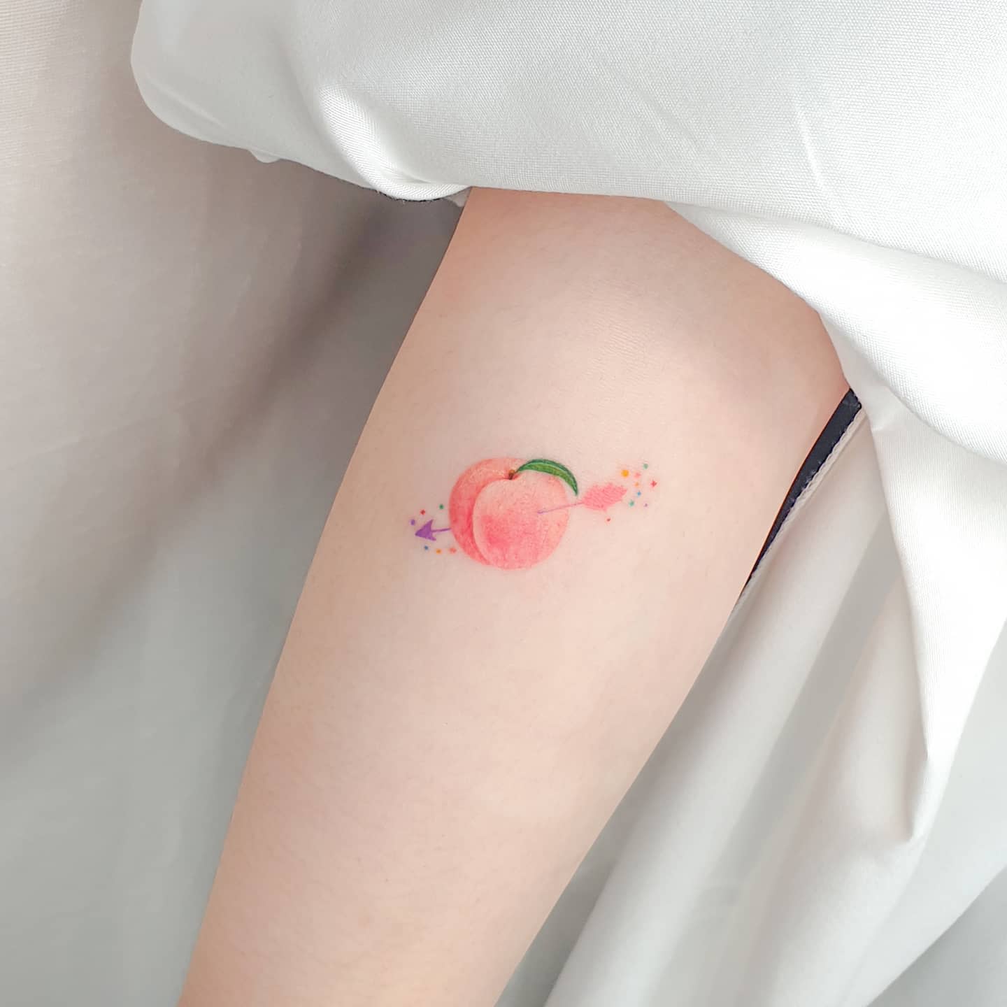 A small peach tattoo with and arrow going through it and colorful sparkles on an arm